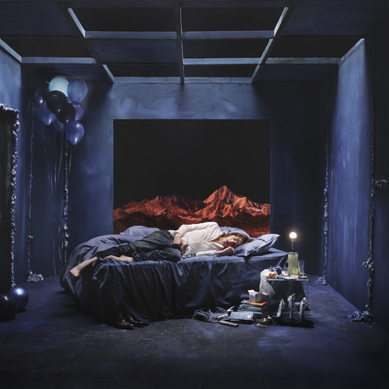 Eine Person liegt voll bekleidet in Jeans und weißem Hemd auf einem ungemachten blauen Bett in einem kargen, bläulich ausgeleuchteten Raum, während im Hintergrund durch eine rechteckige Wandöffnung eine rötlich glühende Gebirgskette unter schwarzem Nachthimmel sichtbar ist.