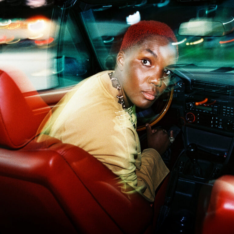 Das Bild zeigt die Musikerin Arlo Parks auf dem Fahrersitz eines Autos bei Nacht. Sie hat kurzes, leuchtend rotes Haar und trägt ein senfgelbes Oberteil sowie eine grobe Gliederkette. Sie blickt über ihre Schulter direkt in die Kamera, während im Hintergrund und durch die Fenster verschwommene Lichtspuren vorbeifahrender Autos und städtischer Beleuchtung zu sehen sind, was eine dynamische Bewegungsunschärfe erzeugt.