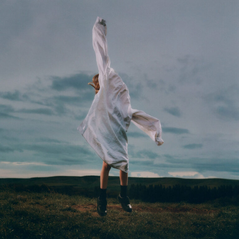 Das quadratische Albumcover zeigt eine Person in Untersicht vor einem weiten, dämmerigen Wolkenhimmel. Die Figur trägt ein langes, weißes Hemd, dessen Ärmel wie Flügel weit nach oben und zur Seite gestreckt sind, während sie vom Boden abhebt. Im unteren Bilddrittel erkennt man eine dunkle, hügelige Graslandschaft mit einer Baumreihe im Hintergrund. Die Lichtstimmung ist kühl und melancholisch.
