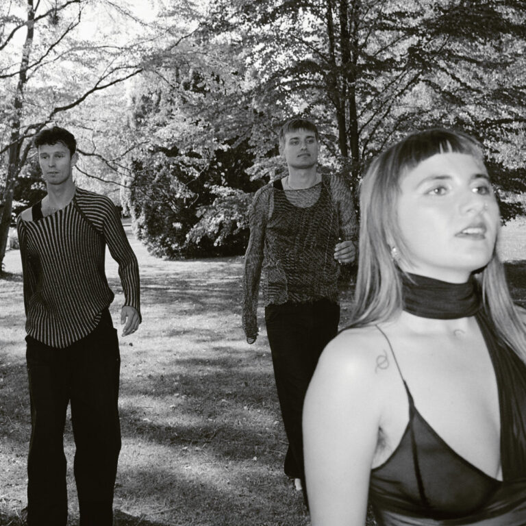 Schwarz-weißes Pressefoto der Band AySay in einem Park. Im Vordergrund rechts steht Sängerin Luna Ersahin mit tätowiertem Symbol auf der Schulter, blickt konzentriert zur Seite. Hinter ihr laufen Aske Døssing Bendixen und Carl West Hosbond in gemusterten, teils transparenten Oberteilen über eine Wiese.