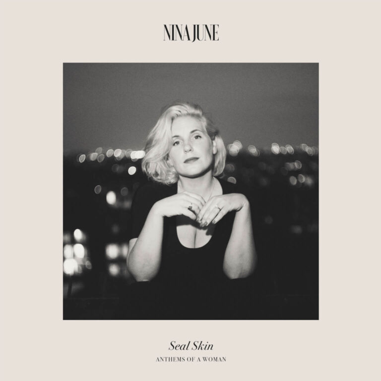 Ein quadratisches Albumcover in Schwarz-Weiß, das eine blondierte Frau mit mittellangem Haar zeigt. Sie trägt ein dunkles Oberteil mit tiefem Ausschnitt und verschränkt die Hände vor der Brust, wobei Ringe an ihren Fingern sichtbar sind. Der Hintergrund ist unscharf und zeigt Lichter einer nächtlichen Stadt in Bokeh-Optik. Der Name „NINA JUNE“ steht oben zentriert, unten befinden sich die Titel „Seal Skin“ und „ANTHEMS OF A WOMAN“.