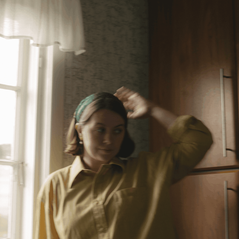 Fotografie einer jungen Frau mit dunklen Haaren und grünem Haarband, die in einer Küche vor einem Fenster steht. Sie trägt ein gelbes Hemd und hält eine Hand nachdenklich am Hinterkopf, während ihr Blick gesenkt ist. Das Licht fällt weich durch das Fenster im Hintergrund.