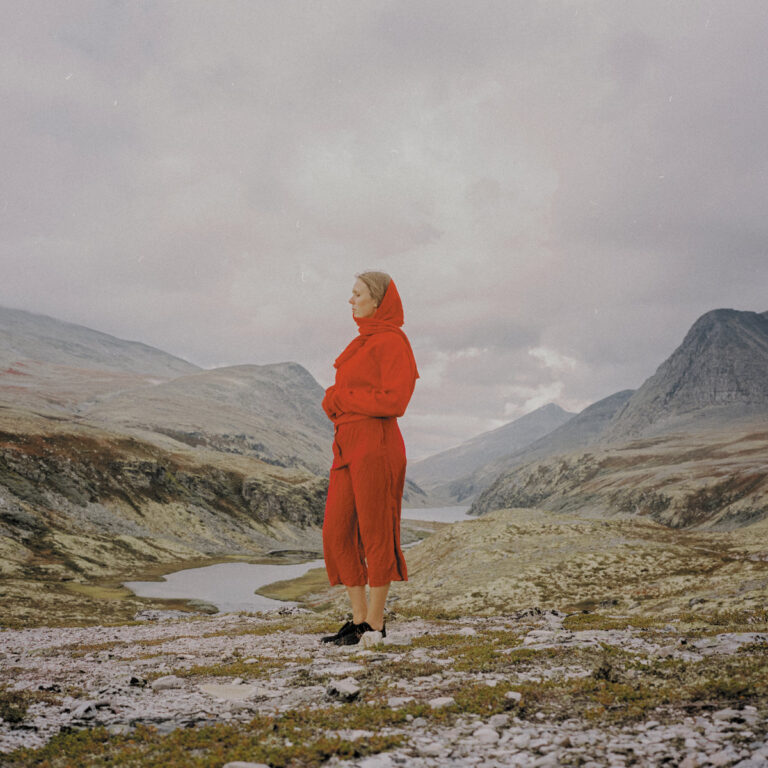 Eine Frau steht im Profil in einer kargen, weiten Gebirgslandschaft Norwegens unter einem wolkenverhangenen Himmel. Sie trägt ein leuchtend rotes Gewand mit Kapuze, das einen starken Kontrast zu den gedämpften Erdfarben der Felsen, Moose und kleinen Seen im Hintergrund bildet.