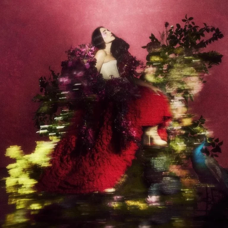 Porträt von Jessie Ware, die in einem opulenten roten Kleid und weißen Plateau-Absätzen inmitten einer dichten, farbenfrohen Blumenlandschaft posiert, während ein Pfau im Vordergrund steht und der Hintergrund in tiefem Magenta erstrahlt.