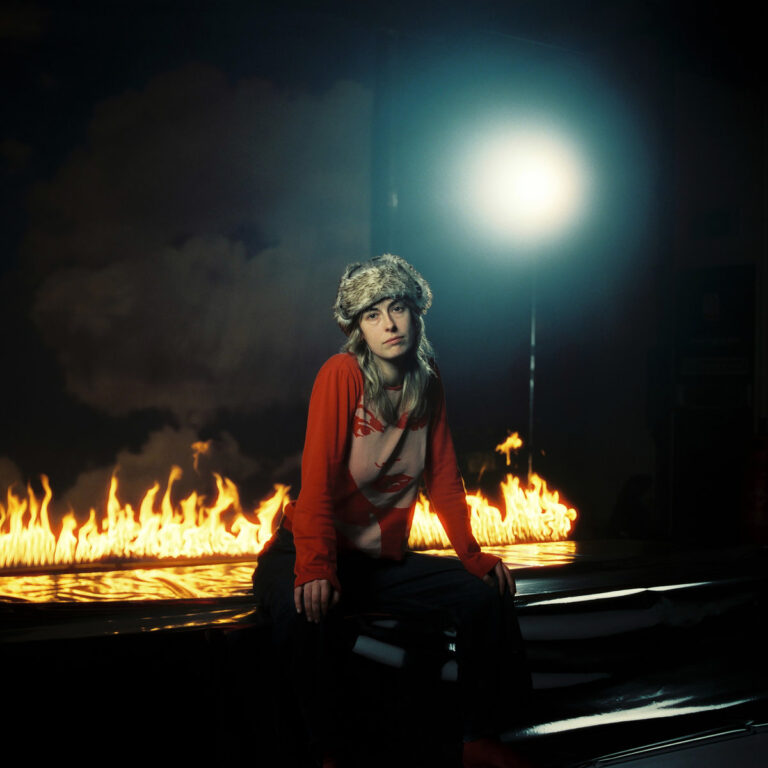 Das quadratische Albumcover zeigt Melanie Baker frontal mittig sitzend auf einer dunklen, reflektierenden Oberfläche. Sie trägt eine helle Pelzmütze und ein langärmliges rotes Oberteil, auf dem ein stilisierter Aufdruck eines Gesichts mit betonten Lippen zu sehen ist. Direkt hinter ihr brennt eine durchgehende Linie aus gelben und orangefarbenen Flammen auf dem Boden. Der Hintergrund ist dunkel und rauchig, während von oben rechts ein starker, bläulich-weißer Lichtkegel auf sie herabscheint und eine künstliche Studioatmosphäre erzeugt.