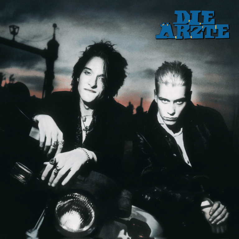 Das quadratische Bild zeigt das Albumcover der Band Die Ärzte. Zwei junge Männer blicken mit ernstem, fast herausforderndem Ausdruck direkt in die Kamera. Sie tragen dunkle Kleidung und Lederjacken. Der Hintergrund ist in ein düsteres, rötliches Dämmerlicht getaucht, das eine vage Silhouette einer Stadt oder eines Friedhofs erahnen lässt. Oben rechts steht in großen, blauen Buchstaben der Bandname „DIE ÄRZTE“.