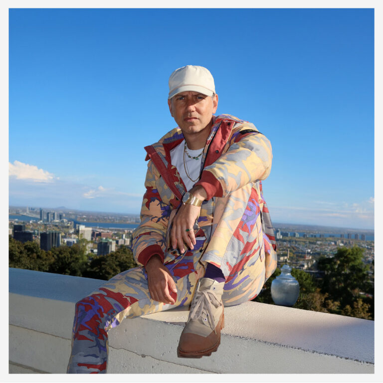 Ein Mann sitzt auf einer hellen Mauer vor einer weiten Stadtlandschaft unter blauem Himmel. Er trägt einen auffälligen Anzug mit Camouflage-Muster in Beige, Rot, Blau und Gelb sowie eine helle Kappe. Er blickt mit ernstem, fokussiertem Ausdruck direkt in die Kamera, die rechte Hand locker im Schoß, die linke Hand stützt sich auf dem Oberschenkel ab.