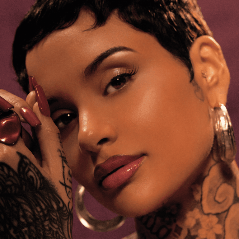 Nahaufnahme des Gesichts von Kehlani vor einem violetten Hintergrund. Die Künstlerin hat kurzes, dunkles Haar und trägt große, silberne Creolen sowie auffällige Tätowierungen am Hals und an den Händen. Ihr Blick ist direkt und intensiv in die Kamera gerichtet, während eine tätowierte Hand mit langen, rötlich lackierten Fingernägeln sanft ihr Gesicht berührt.