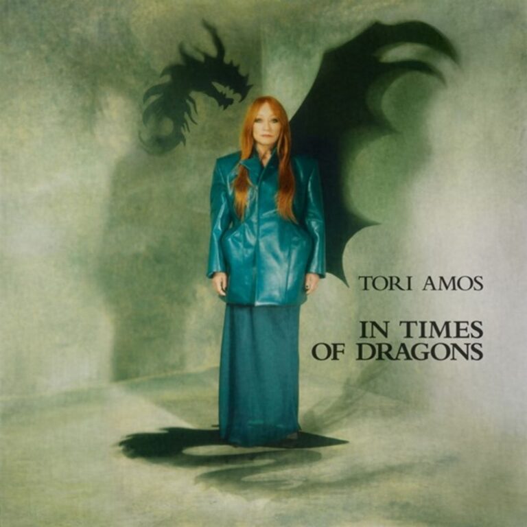 Porträtaufnahme von Tori Amos in einem smaragdgrünen Lederoutfit vor einer strukturierten Wand; hinter ihr zeichnet sich ein großer Schatten in Form eines Drachen mit gespreizten Flügeln ab.