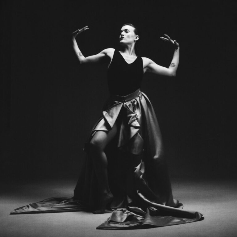 Schwarz-weiß-Fotografie einer Künstlerin in einer theatralischen Pose. Sie trägt ein dunkles, ärmelloses Oberteil und einen weit schwingenden, bodenlangen Rock, der sich stoffreich auf dem Boden ausbreitet. Ihre Arme sind angewinkelt nach oben erhoben, die Hände in einer grazilen Geste gespreizt, während sie den Kopf leicht in den Nacken legt und zur Seite blickt.