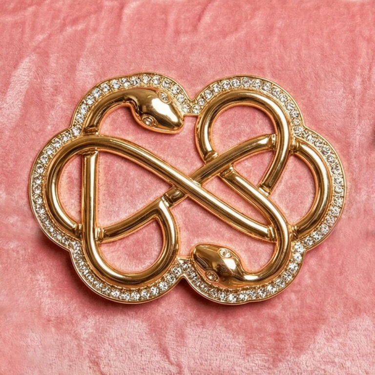 Ein quadratisches Bild zeigt eine goldene, mit kleinen weißen Steinen besetzte Brosche in Form einer liegenden Acht oder eines Unendlichkeitszeichens, dessen Enden als Schlangenköpfe gestaltet sind. Das Schmuckstück liegt zentral auf einem strukturierten, rosa-violetten Hintergrund, der an Stoff oder eine grob verputzte Wand erinnert.