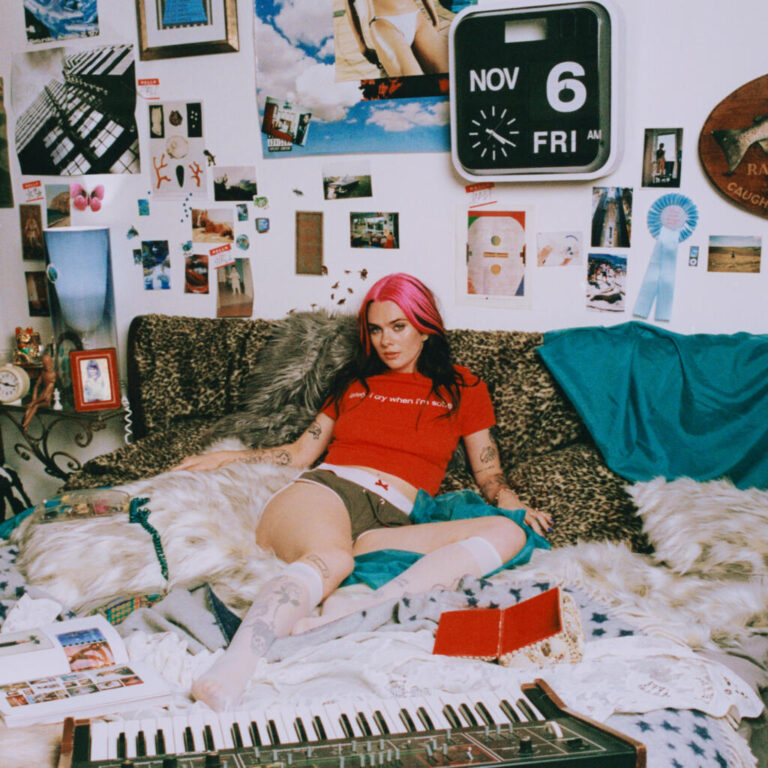 Das quadratische Bild zeigt eine junge Frau mit leuchtend pink gefärbtem Haar, die mittig auf einem unordentlichen Sofa oder Bett sitzt. Sie trägt ein rotes T-Shirt und kurze dunkle Hosen. Die Wand hinter ihr ist überladen mit Postern, Fotos, Zetteln und einer großen, quadratischen Bahnhofsuhr, die Freitag, den 6. November anzeigt. Im Vordergrund liegt ein elektronisches Keyboard auf einer weißen Decke, umgeben von verstreuten Papieren. Die Atmosphäre wirkt wie ein privates, kreativ-chaotisches Jugendzimmer mit einer leicht nostalgischen, analogen Ästhetik.