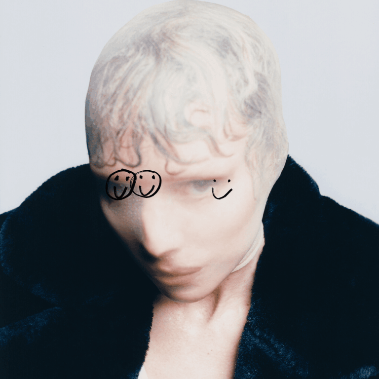 Porträtaufnahme der Künstlerin Lykke Li, die ihren Kopf mit einer hellen Nylonmaske verhüllt hat, auf der im Augenbereich zwei einfache, stilisierte lachende Gesichter und ein kleiner Bogen gezeichnet sind. Sie trägt einen dunklen, strukturierten Pelzmantel oder eine Jacke vor einem neutralen, hellen Hintergrund.