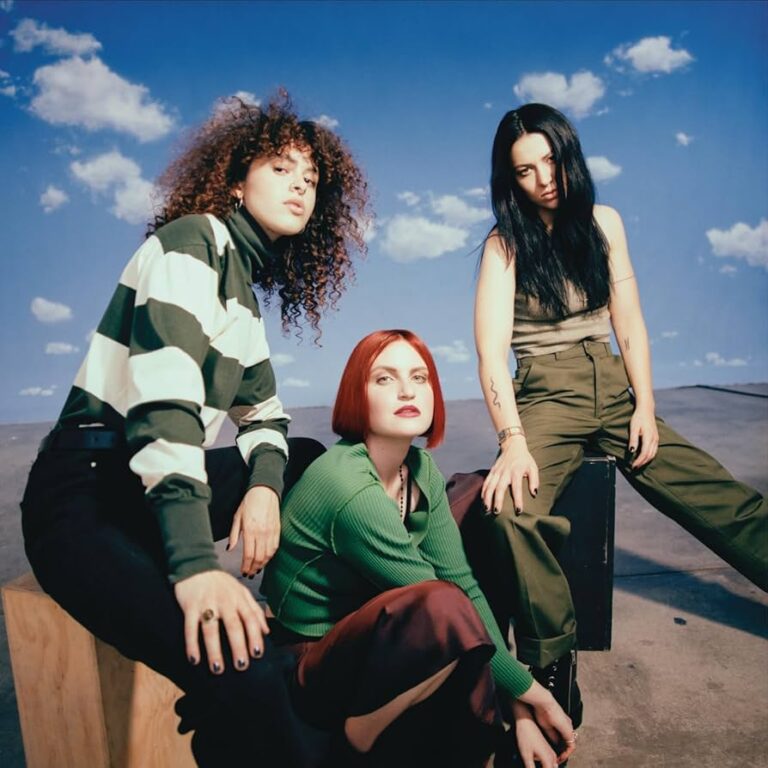 Das Bild zeigt das Trio MUNA – Josette Maskin, Katie Gavin und Naomi McPherson – in einer leicht untersichtigen Perspektive vor einem strahlend blauen Himmel mit vereinzelten weißen Wolken. Die Musikerinnen tragen Outfits in gedeckten Tönen wie Dunkelgrün, Braun und gestreift, während sie auf hölzernen Boxen in einer kargen, fast wüstenartigen Landschaft sitzen.