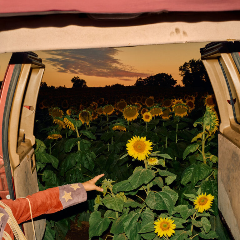 Das quadratische Bild zeigt den Blick aus dem geöffneten Heck eines beige-braunen Lieferwagens auf ein Feld voller Sonnenblumen in der Abenddämmerung. Im linken Vordergrund ragt ein Arm ins Bild, der eine rotbraune Wildlederjacke mit hellen Fransen und zwei aufgenähten Sternen trägt. Die Hand ist ausgestreckt und scheint in Richtung der Sonnenblumen zu greifen. Der Himmel im Hintergrund ist in warmen Orange- und Gelbtönen gefärbt, während einzelne dunkle Wolkenfetzen und Silhouetten von Bäumen am Horizont zu sehen sind.