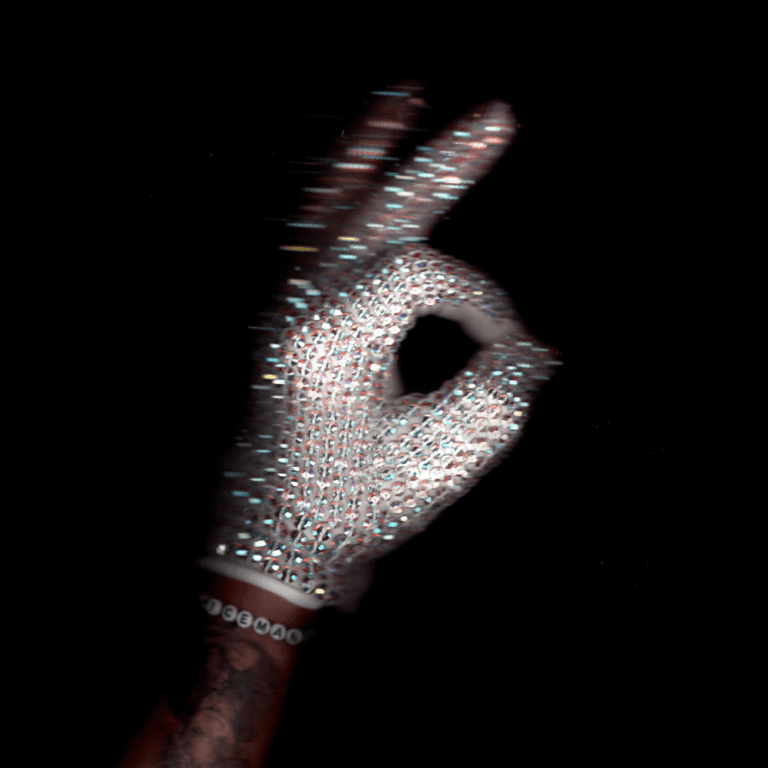 Hand im glitzernden, diamantbesetzten Handschuh vor schwarzem Hintergrund formt das OK-Zeichen mit leichtem Bewegungsunschärfe-Effekt.