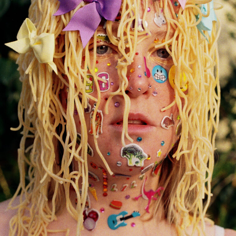 Close-up eines Gesichts mit Nudeln als Haare und vielen bunten Stickern auf der Haut.