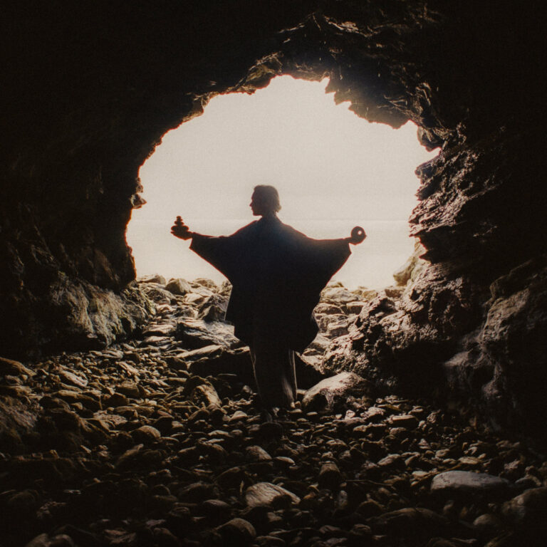 Silhouettierte Person mit ausgebreiteten Armen im hellen Ausgang einer dunklen, felsigen Höhle.