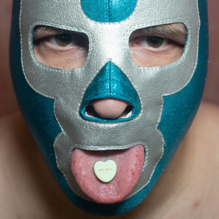 Close-up eines Gesichts mit blau-silberner Wrestling-Maske und einem Herz-Bonbon auf der herausgestreckten Zunge.