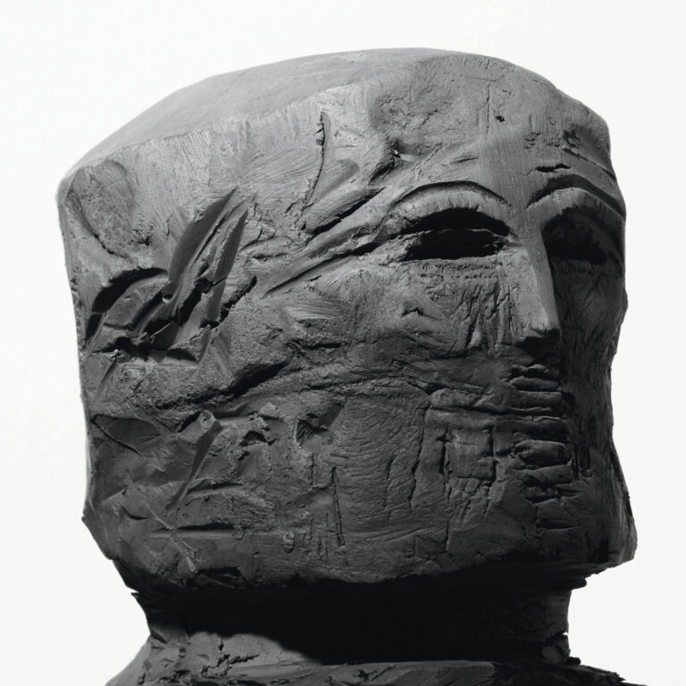 Eine grob geformte, dunkle Tonskulptur eines menschlichen Kopfes vor weißem Hintergrund.
