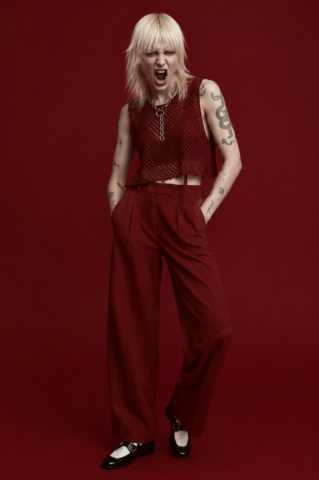 Eine junge Frau mit hellblondem Haar steht in einem vollständig monochromen Studio in tiefem Bordeauxrot und blickt mit offenem Mund und entschlossener Aggression direkt in die Kamera. Nur ihre helle Haut, das Haar und das schwarze Schuhwerk heben sich von der exakt einfarbigen Umgebung und Kleidung ab, während markante Tätowierungen den rebellischen Charakter unterstreichen.