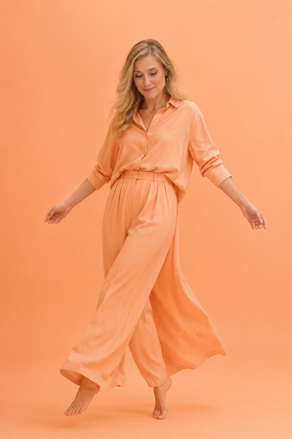 Eine Frau bewegt sich scheinbar schwerelos durch einen vollständig monochromen Raum in hellem Apricot, während ihr Blick mit einem dezenten Lächeln zum Boden gerichtet ist. Das fließende Outfit, die weiten Stoffbahnen und die sanfte Körperhaltung verleihen der Szene eine leichte, unbeschwerte Eleganz.