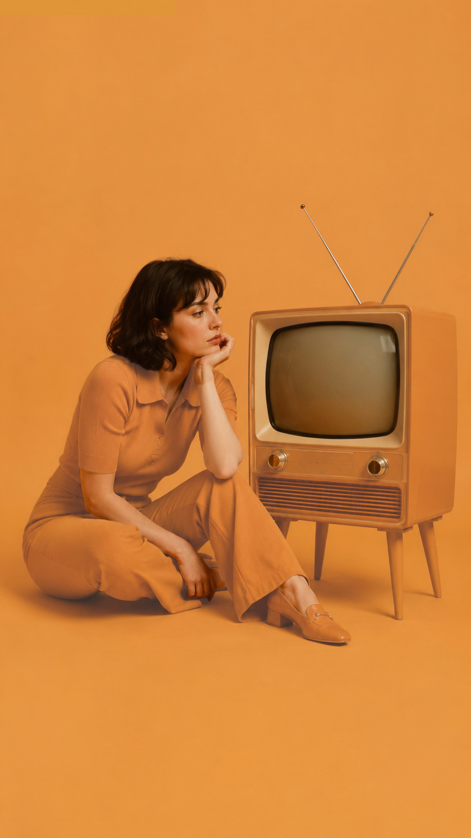 Eine Frau sitzt nachdenklich neben einem Retro-Fernseher in einem vollständig monochromen Raum in nostalgischem Orange. Nur Haut, Haare und der Fernseher heben sich farblich vom exakt einheitlichen Hintergrund und der Kleidung ab.