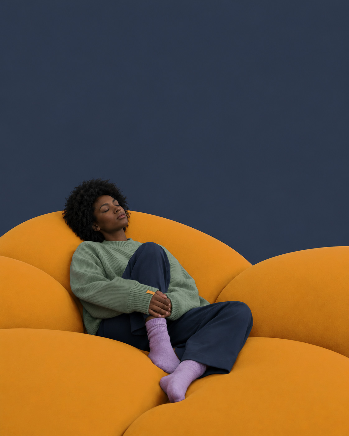 Eine Frau mit geschlossenen Augen versinkt in einem übergroßen Sofa und verliert sich in stillen Gedanken. Die weiten Flächen und die ruhige Farbwelt fangen die entschleunigte Atmosphäre eines einsamen Sonntagnachmittags ein.