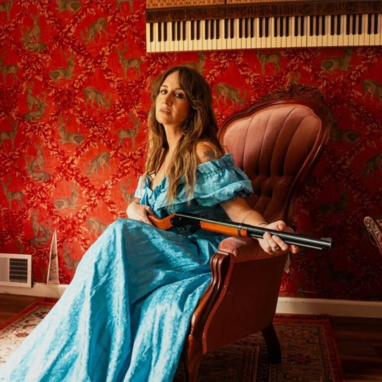 Margo Price sitzt im blauen Kleid mit einem Gewehr auf einem Sessel vor roter Tapete.