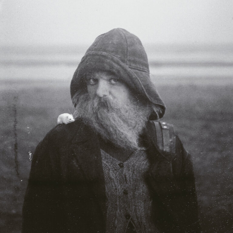 Schwarz-Weiß-Fotografie eines bärtigen Einsiedlers mit Kapuze und einer kleinen Maus auf der Schulter.