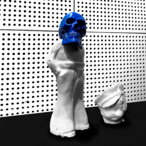 Ein Gipsabdruck mit blauem Totenkopf vor einer sterilen Lochwand.