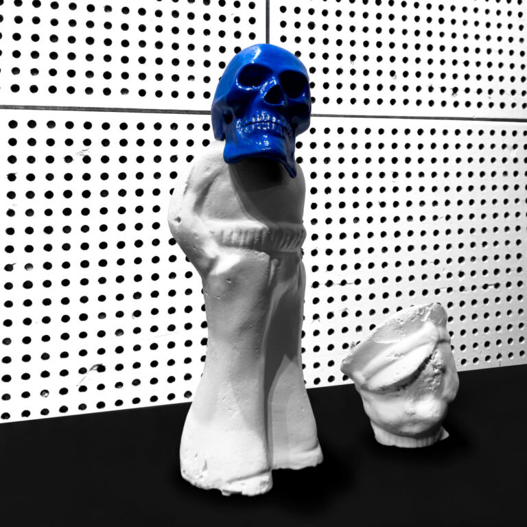 Ein Gipsabdruck mit blauem Totenkopf vor einer sterilen Lochwand.