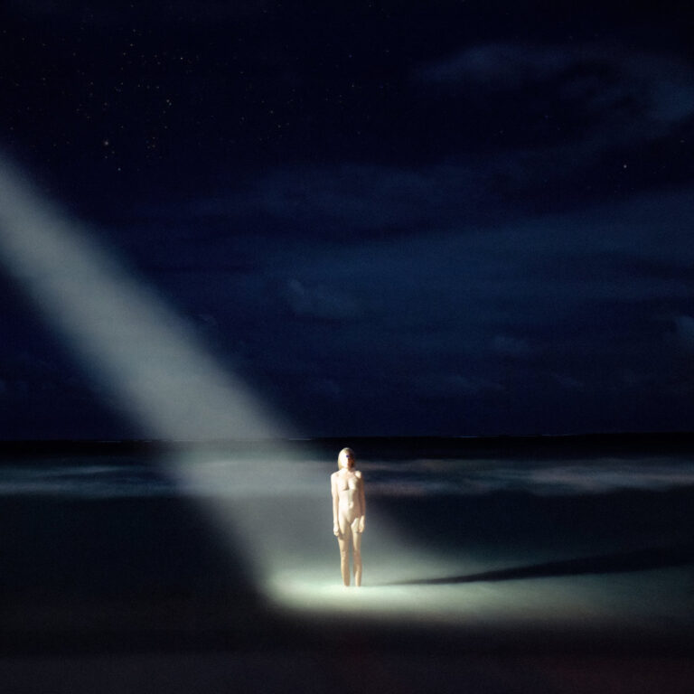 Eine nackte Gestalt steht im Scheinwerferkegel am nächtlichen Strand.