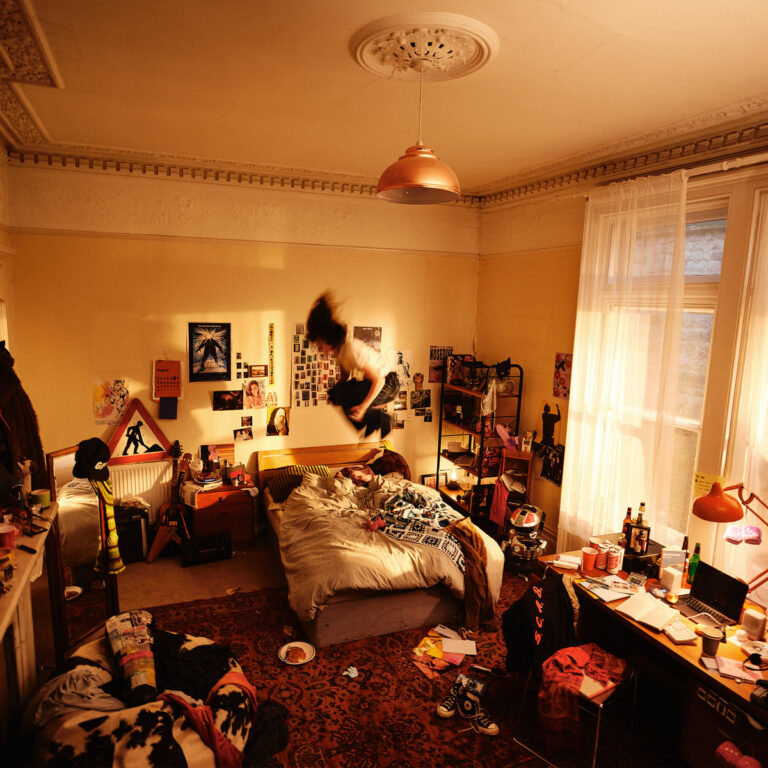 Eine Person springt in einem sonnendurchfluteten, unaufgeräumten Jugendzimmer dynamisch auf ein Bett.