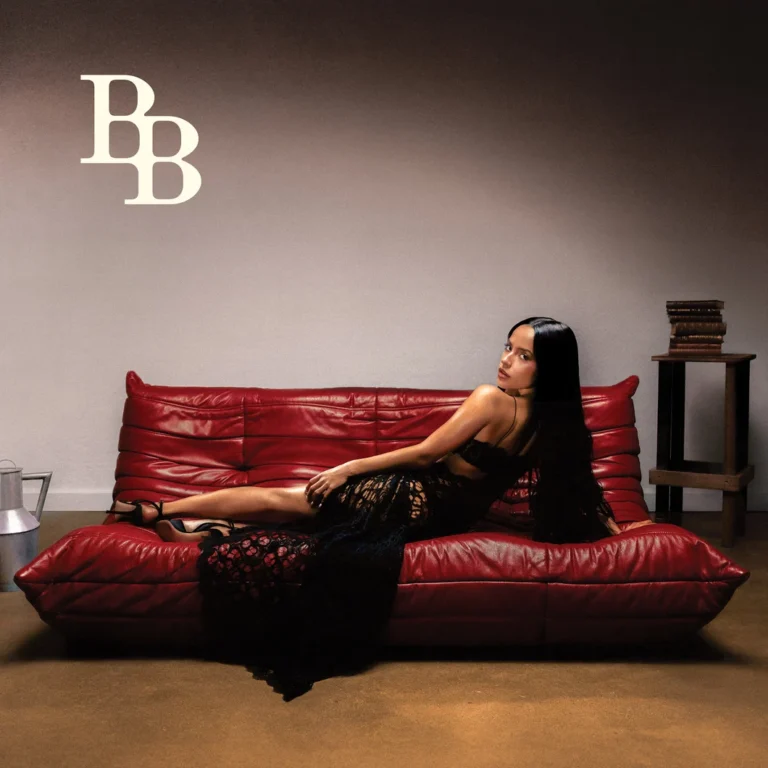 Becky G liegt in einem schwarzen Spitzenkleid auf einem roten Ledersofa.