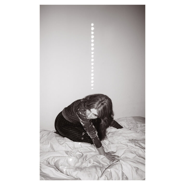 Schwarz-Weiß-Foto von Julia Holter auf einem zerknüllten Bett mit Lichtreflexen an der Wand.