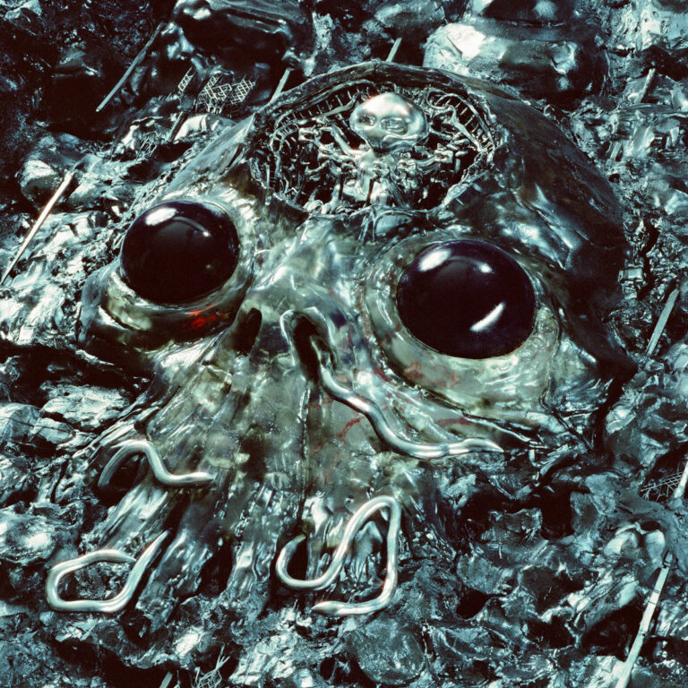 Glänzender Totenkopf aus geschmolzenem Metall mit dunklen Kugelaugen und Tentakeln inmitten von Schrott.