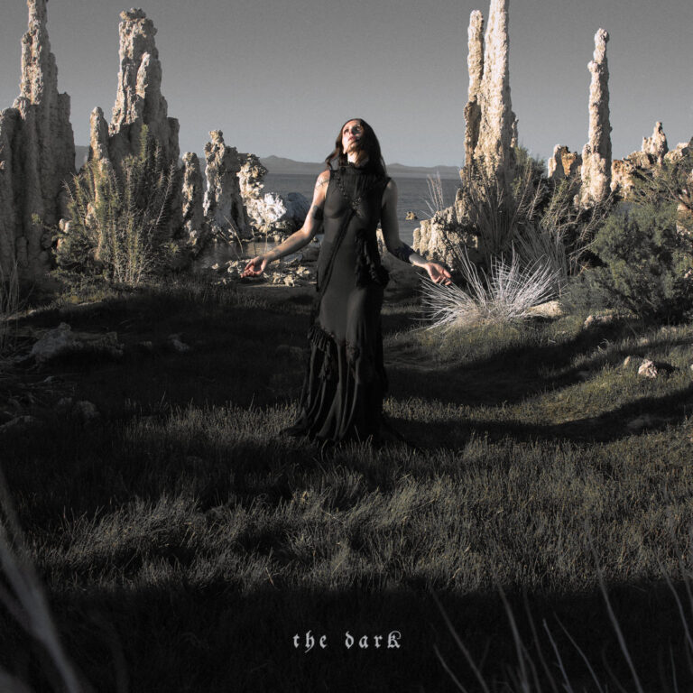 Chelsea Wolfe steht in einem schwarzen Kleid in einer steinigen Landschaft.