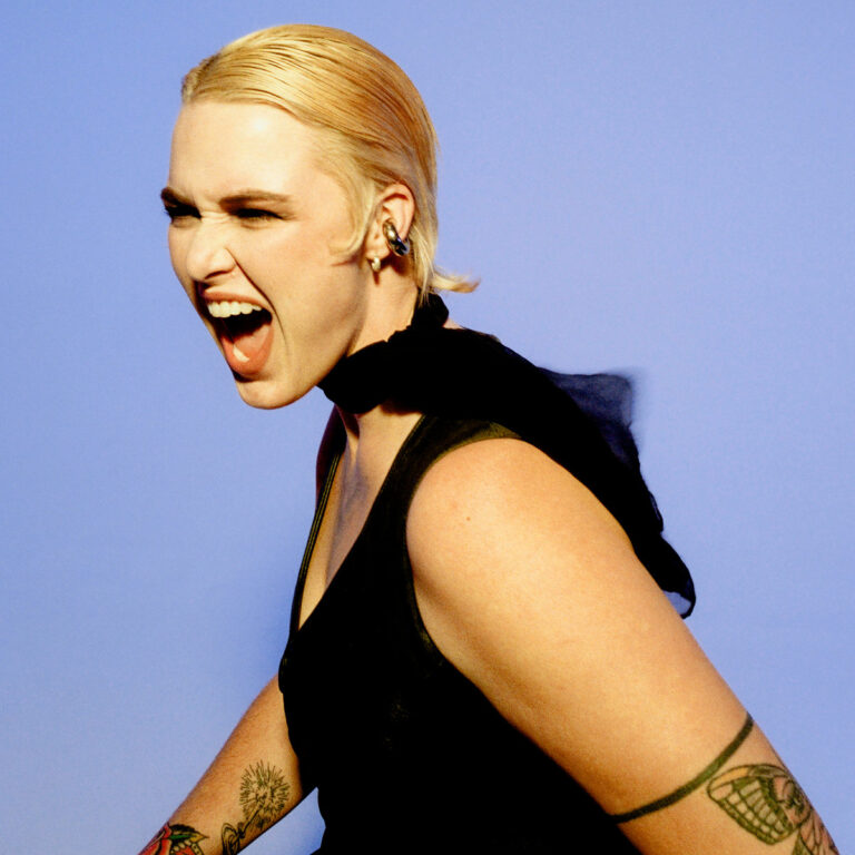 Eine blonde Frau mit Tattoos schreit kraftvoll im Profil vor blauem Hintergrund.