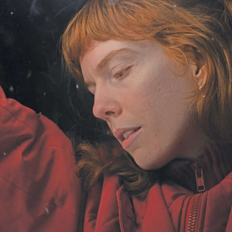 Nahaufnahme von Elanor Moss mit roter Jacke im Profil vor dunklem Hintergrund.