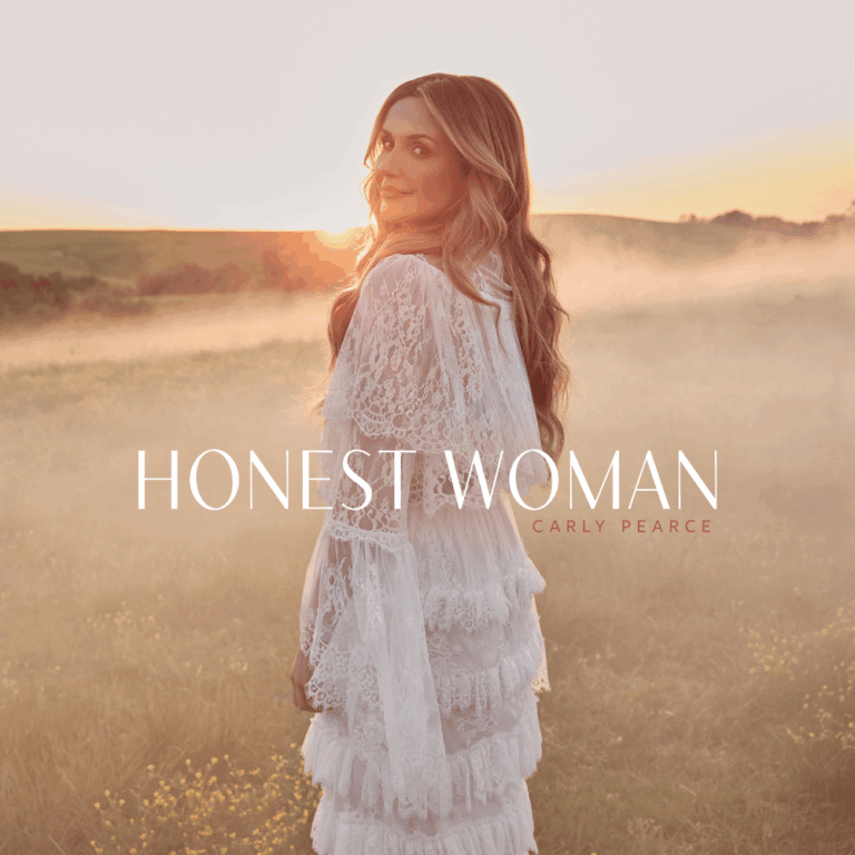 Carly Pearce steht in einem weißen Spitzenkleid auf einem nebligen Feld im Abendlicht.