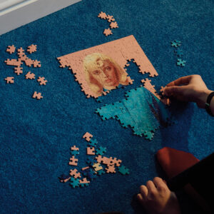 Eine Hand setzt auf blauem Teppich ein Puzzle zusammen, das ein unvollständiges Porträt einer blonden Frau zeigt.