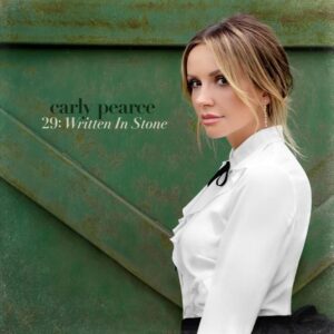 Carly Pearce blickt im Profil in einem weißen Hemd mit schwarzer Schleife vor einer grünen Holzscheune in die Kamera.