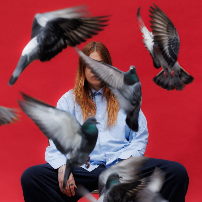 Die Musikerin Leila sitzt vor rotem Hintergrund in einem hellblauen Hemd, während fliegende Tauben ihr Gesicht vernebeln.