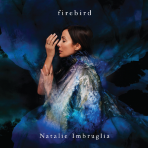 Natalie Imbruglia posiert im Profil vor dunkelblauem Hintergrund in einem blau strukturierten Tüllkleid mit einer Hand am Kopf.